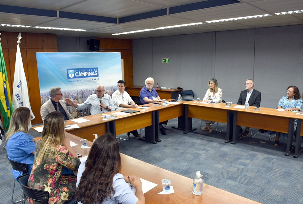 foto mostra pessoas sentadas em mesas, no formato de U, em uma sala da prefeitura de campinas