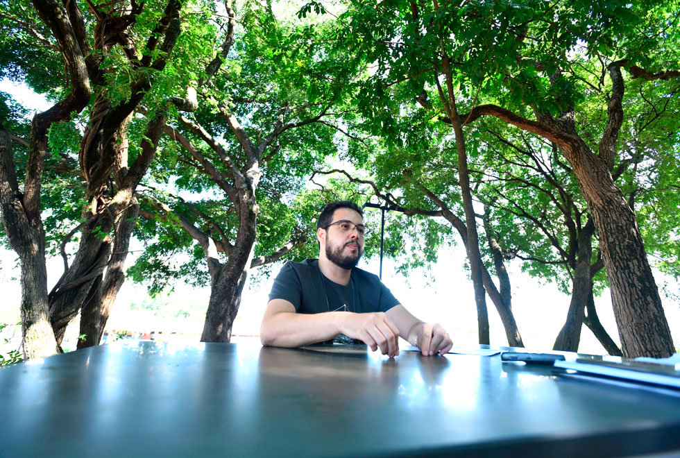 audiodescrição: fotografia colorida mostra um homem gesticulando enquanto fala. ele está sentado em um banco em frente a uma mesa ao ar livre. ao fundo há árvores e um dia ensolarado.