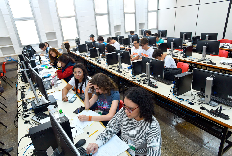 audiodescrição: fotografia colorida mostra estudantes em quatro fileiras de mesas sentados a frente de computadores, em uma sala onde acontece uma maratona de programação. na primeira fileira, estão em maioria meninas. nas outras, a maioria é de meninos. 