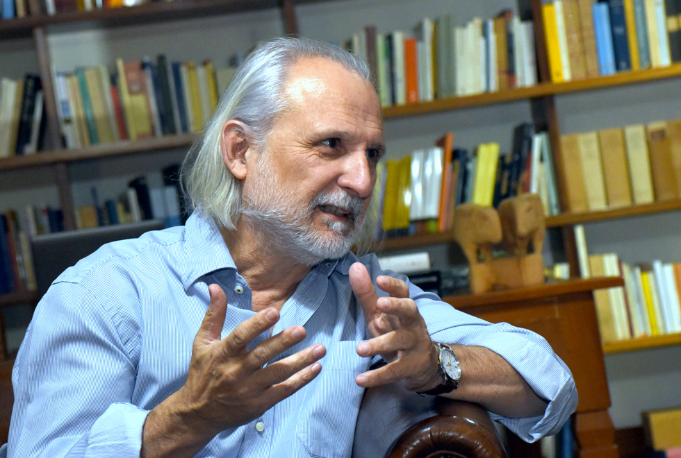 foto mostra o professor alcir pécora em uma biblioteca. Ele veste camisa azul e tem cabelos na altura dos ombros