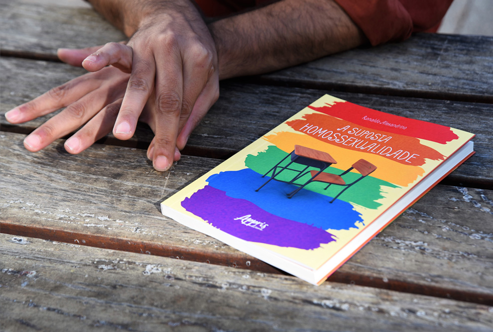 audiodescrição: fotografia colorida do livro "a suposta homossexualidade"; o livro está em cima de uma mesa e ao lado estão as mãos do autor