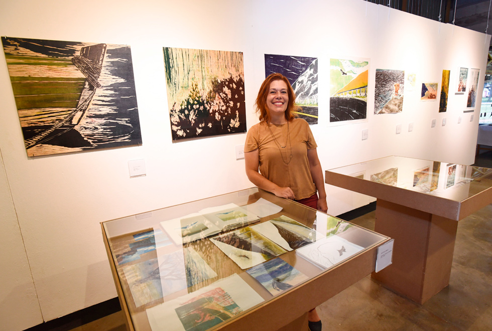 Audiodescrição: fotografia colorida mostra a mestranda Ana Paula de Melo sorrindo, em pé em meio a sua exposição, que conta com obras penduradas na parede e duas vitrines de vidro no chão.