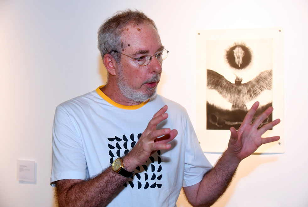 Audiodescrição: fotografia colorida professor Sérgio gesticulando enquanto dá entrevista. Ao fundo, na parede da galeria, há um trabalho artístico de seu orientando.