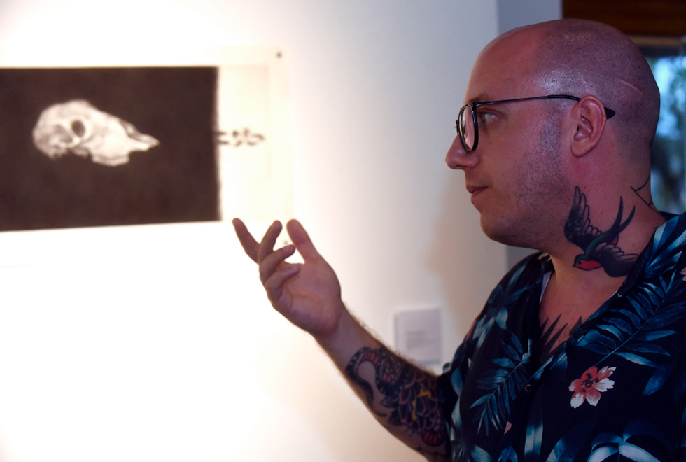 Audiodescrição: fotografia colorida mostra estudante de mestrado André Berger de perfil gesticulando. Ao fundo, há uma de seus trabalhos, em preto e branco.