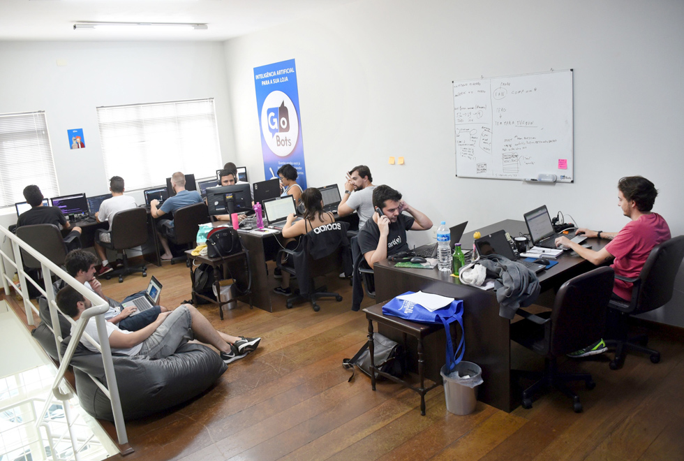 foto mostra o escritório da empresa gobots com funcionários sentados em escrivaninhas trabalhando em laptops e outros sentados em pufes. O ambiente é descontraído