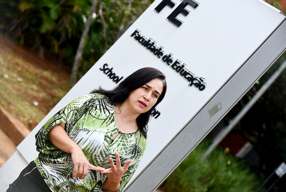 audiodescrição: fotografia colorida da professora Luciane Barbosa em frente à placa de entrada da faculdade de educação da unicamp