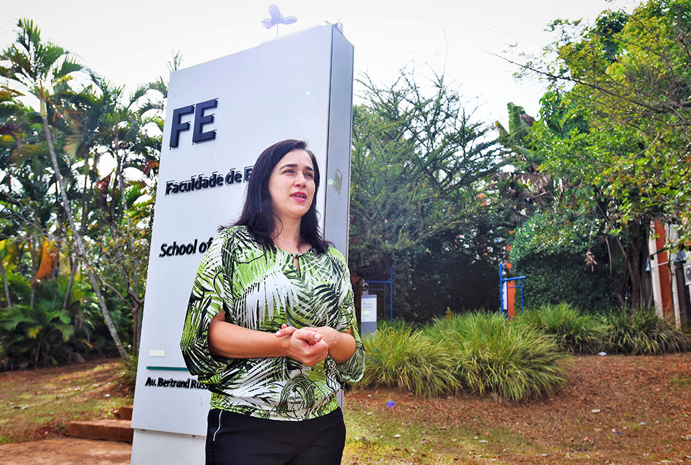 audiodescrição: fotografia colorida da professora Luciane Barbosa em frente à placa de entrada da faculdade de educação da unicamp