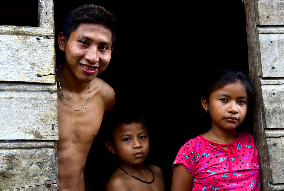 audiodescrição: fotografia colorida de três jovens indígenas posando para foto em frente a uma casa de madeira