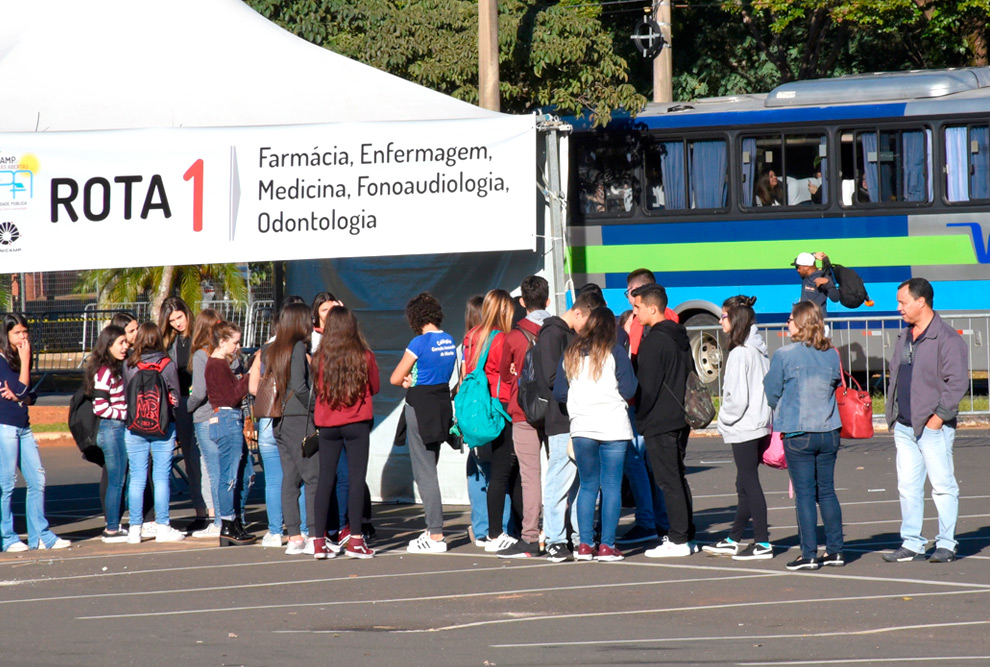 foto mostra alunos em fila em frente a uma barraca com faixa indicando "rota 1"