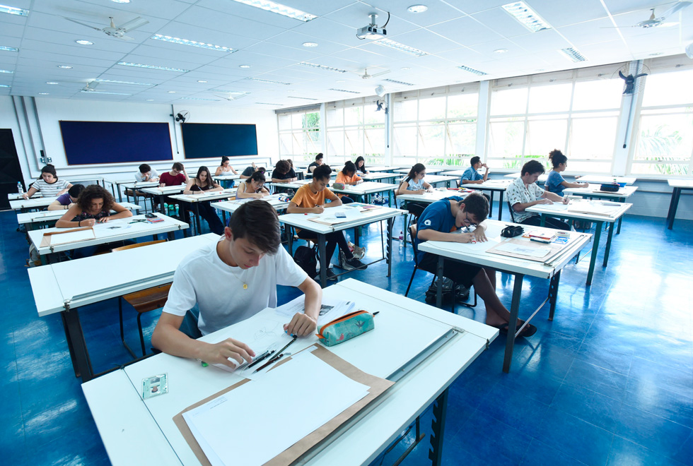 Audiodescrição: fotografia colorida mostra alunos em uma sala de aula, durante a prova de habilidades para ingresso no curso de arquitetura e urbanismo eles estão concentrados e em cada mesa há materiais de desenho.