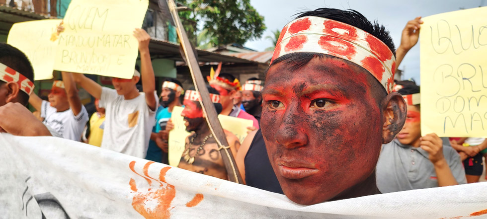 audiodescrição: imagem colorida, em foco manifestante indígena com o rosto pintado e parte de uma faixa aparecendo. (foto:Antonio Scarpinetti)