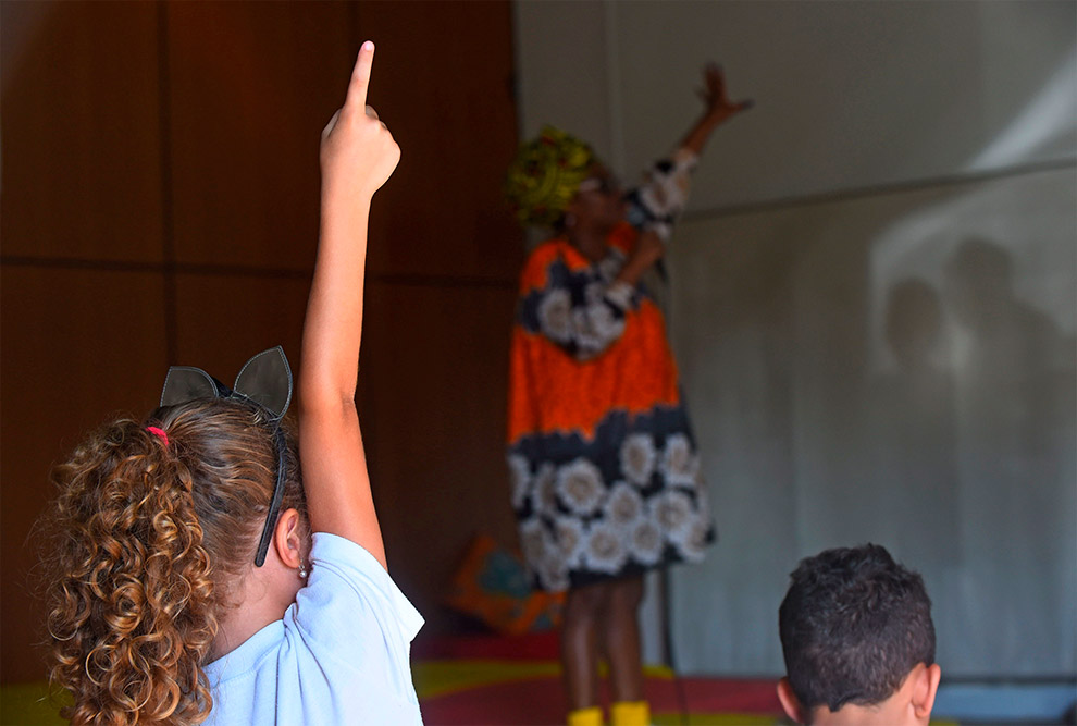 mão de uma criança da plateia levantada em foco, atrás vemos a escritora no palco do evento de contação de histórias