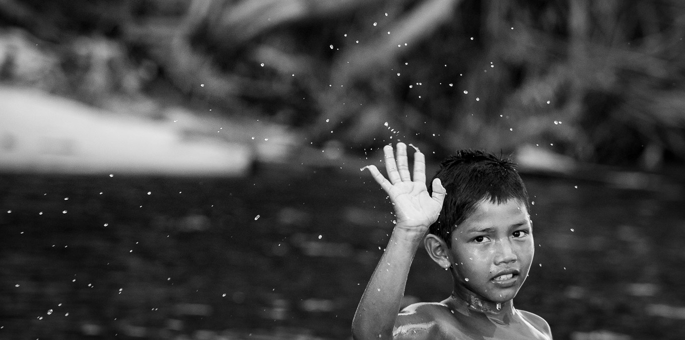 audiodescrição: fotografia em branco e preto de menino indígena brincando no rio enquanto acena para o fotógrafo.