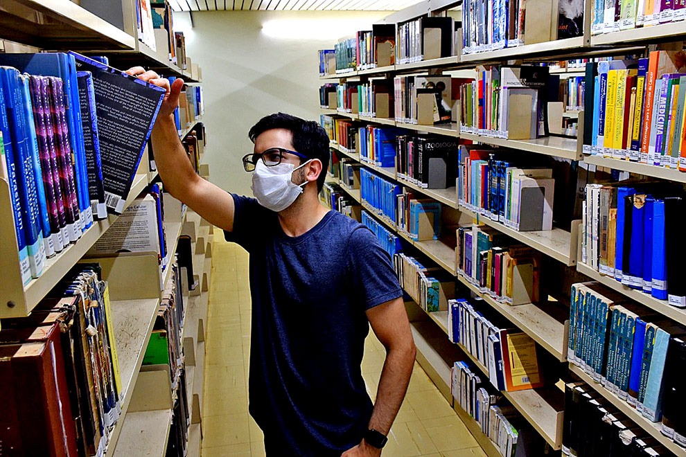 foto mostra estudante de máscara retirando livro de uma estante