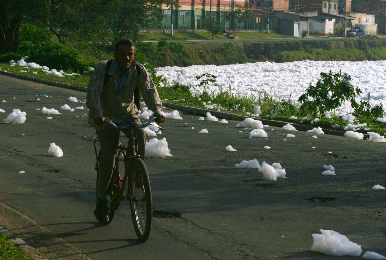 Ciclista passa ao lado de rio coberto por espumas brancas em foto que mostra poluição das águas