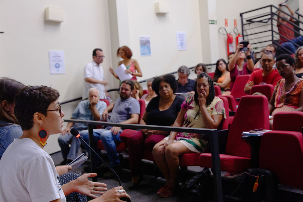 audiodescrição: fotografia colorida de auditório com pessoas assistindo à fala de talita, que está em primeiro plano falando em microfone