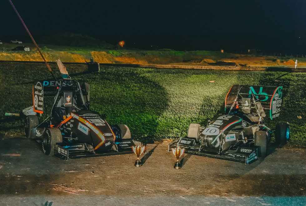 audiodescrição: fotografia colorida de dois carros da E-Racing juto aos trofeus recebidos em competições