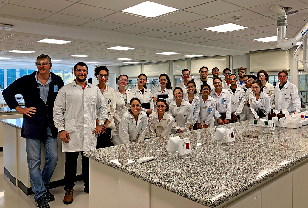 audiodescrição: fotografia colorida mostra diversos estudantes de jaleco branco dentro das instalações da empresa Croda, junto ao professor Edvaldo, da Unicamp. eles estão posando para foto, sorrindo, e em frente há um balcão de laboratório.