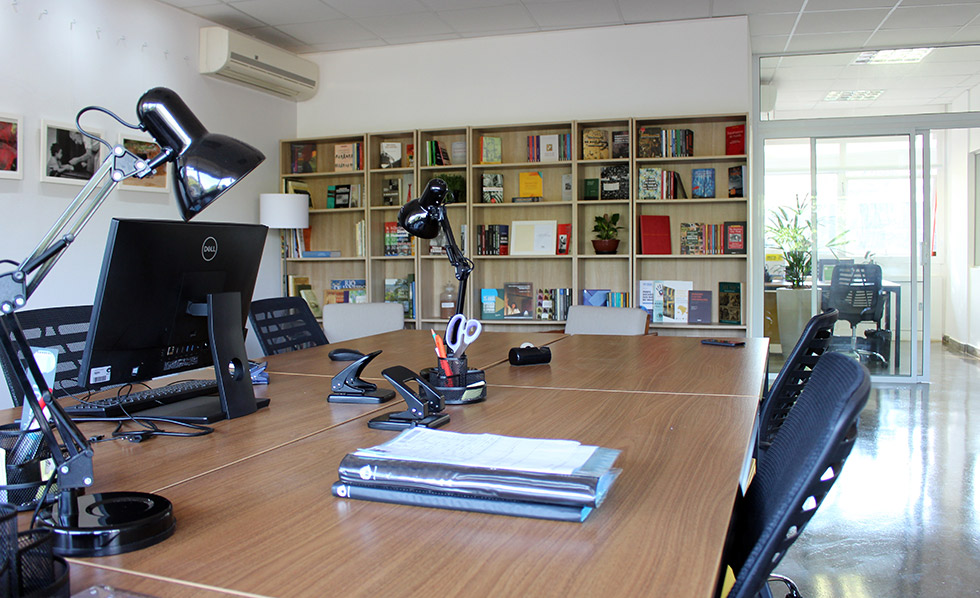 foto mostra uma mesa de trabalho em primeiro plano, com luminárias, e uma estante com livros em segundo plano