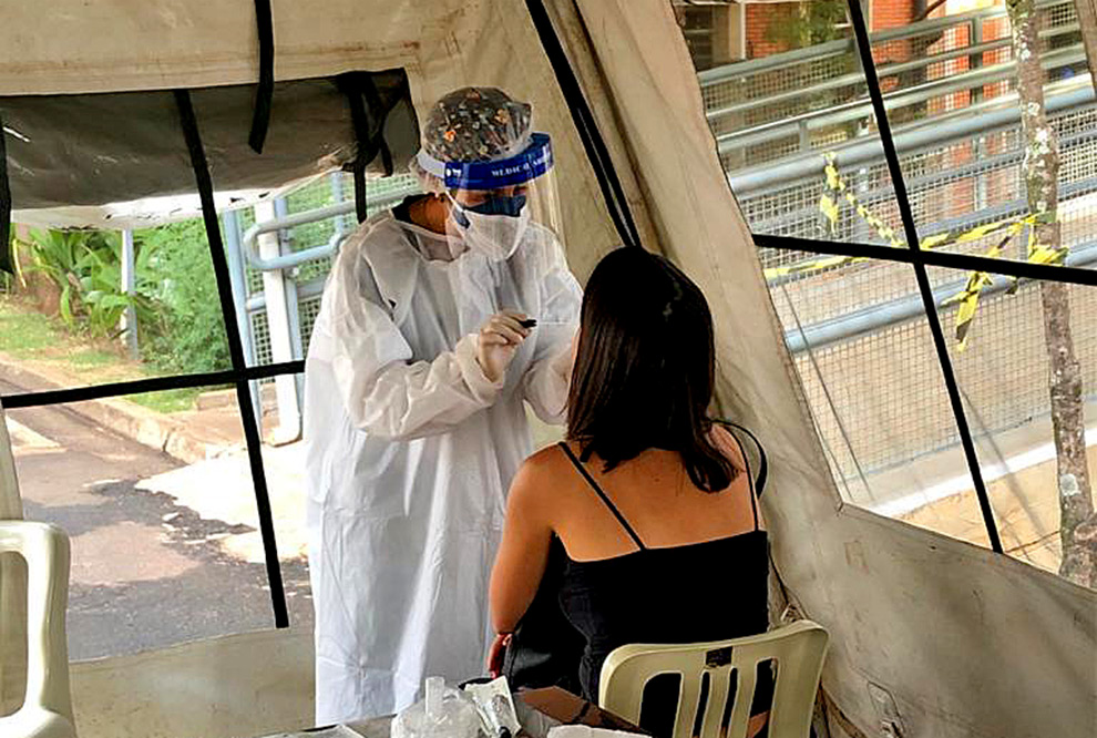 foto mostra um profissional de saúde, paramentado com roupa de proteção, máscara e face shiled, coletando exame de uma moça sentada em uma cadeira