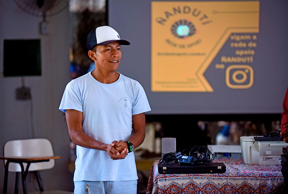 Audiodescrição: fotografia colorida mostra estudante indígena em pé. Ele está falando e atrás dele há uma tela onde está projetado o logo da rede de apoio aos estudantes indígenas.