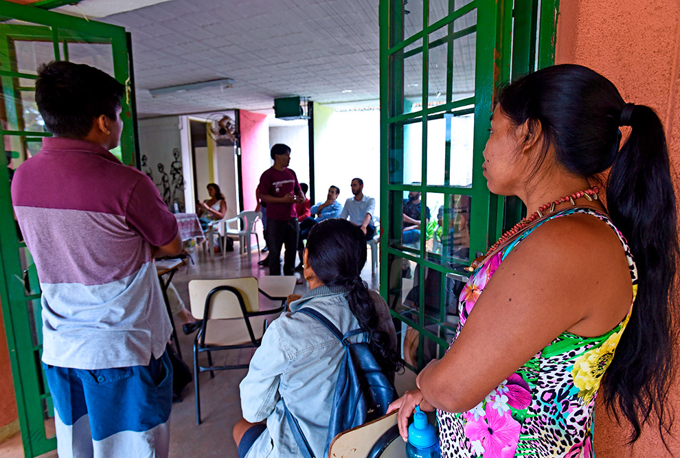 Audiodescrição: fotografia colorida mostra três pessoas de costas olhando para uma reunião que acontece dentro de uma sala