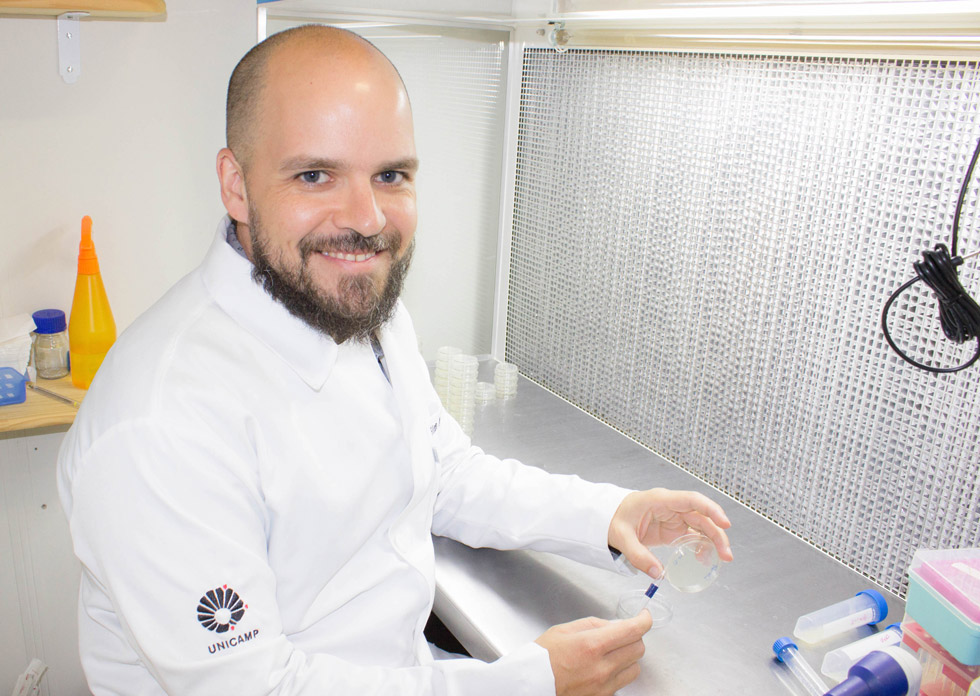 foto mostra marcelo mori em um laboratório mexendo com instrumentos e sorrindo para câmera. ele veste jaleco branco