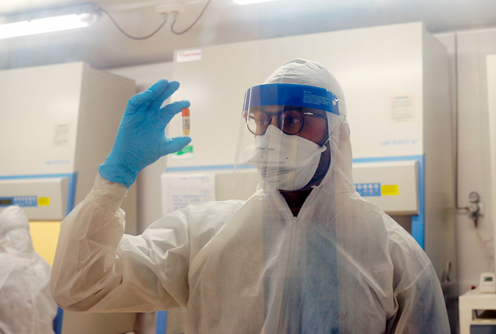 foto mostra pesquisadores dentro de um laboratório, usando roupas de proteção. o que está em primeiro plano segura um frasco com amostra do coronavírus na altura dos olhos