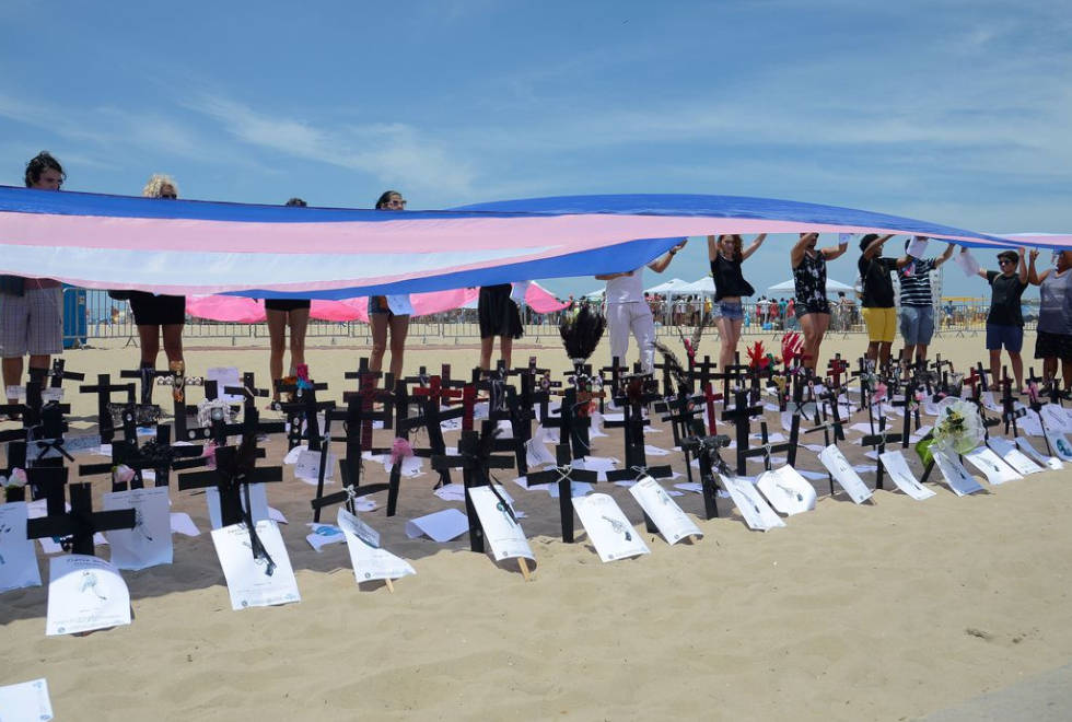 foto mostra uma manifestação em areia de praia com cruzes fincadas na areia cobertas por uma bandeira nas cores azul, branco e rosa, símbolo da comunidade trans