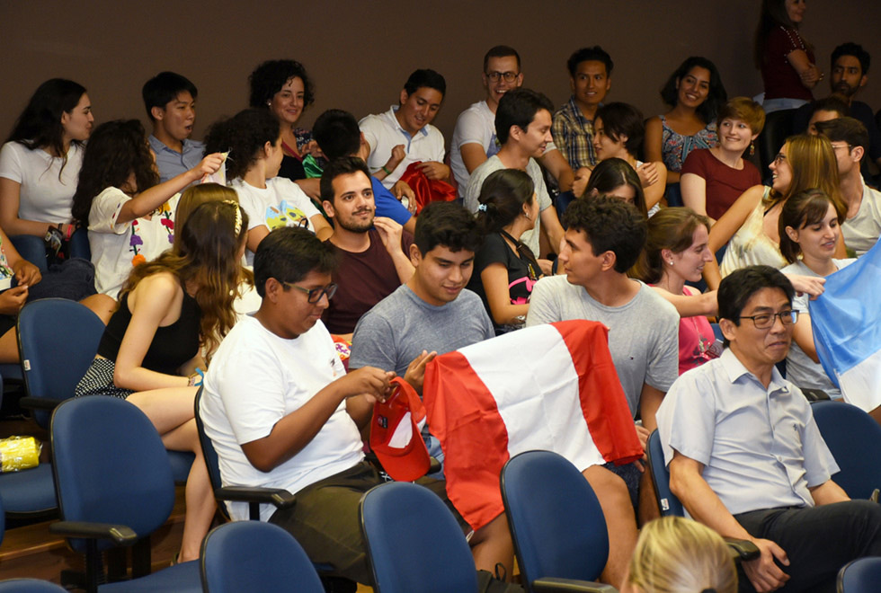 audiodescrição: fotografia colorida de estudantes sentados em auditório; dois deles seguram uma bandeira do peru