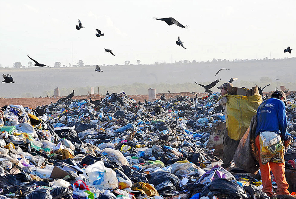 audiodescrição: fotografia colorida de um aterro com lixo; duas pessoas estão trabalhando na seleção