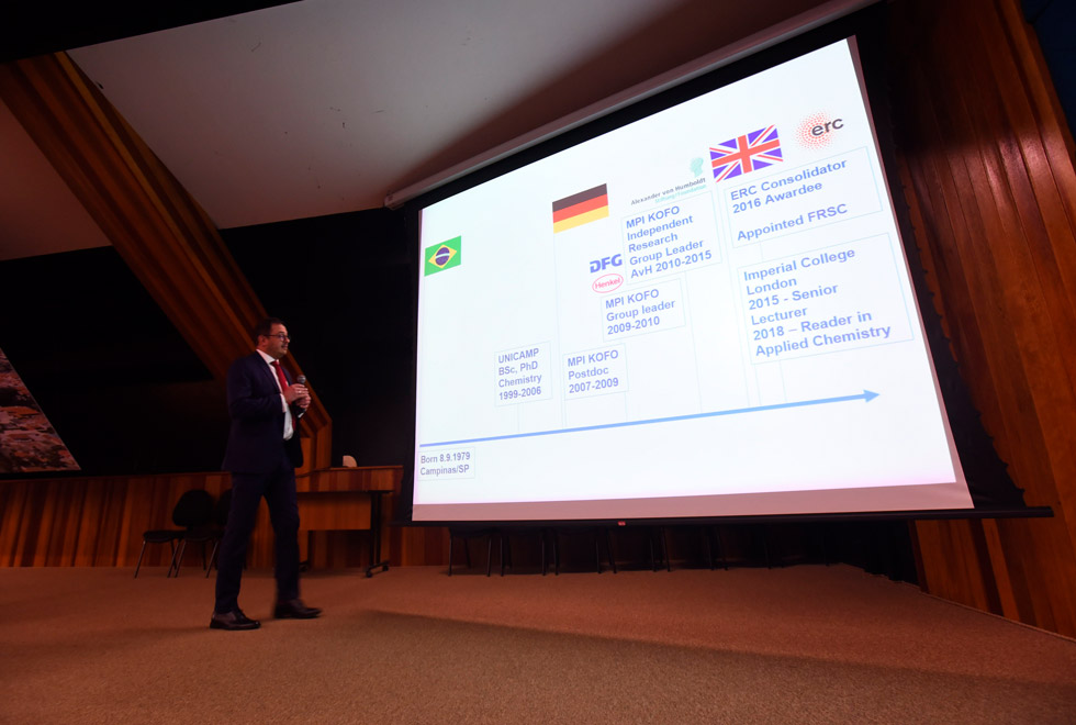 audiodescrição: fotografia colorida mostra professor roberto rinaldi em cima de palco enquanto ministra aula magna. há um slide projetado mostrando a trajetória acadêmica dele