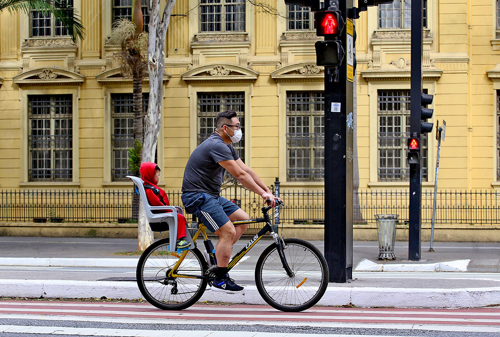 foto mostra homem andando de bicicleta com uma criança na garupa. ambos usam máscaras