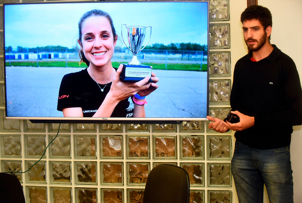 foto mostra renan rodrigues durante uma apresentação em frente a uma tela. a tela mostra uma ex-aluna, iara arromba, segurando um troféu