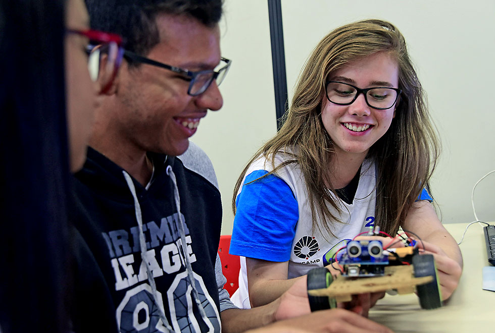 Estudantes observam sobre a mesa e à frente do computador o pequeno robô que é uma estrutura com duas rodas. Dois adolescentes estão sorrindo. Uma menina e um menino.