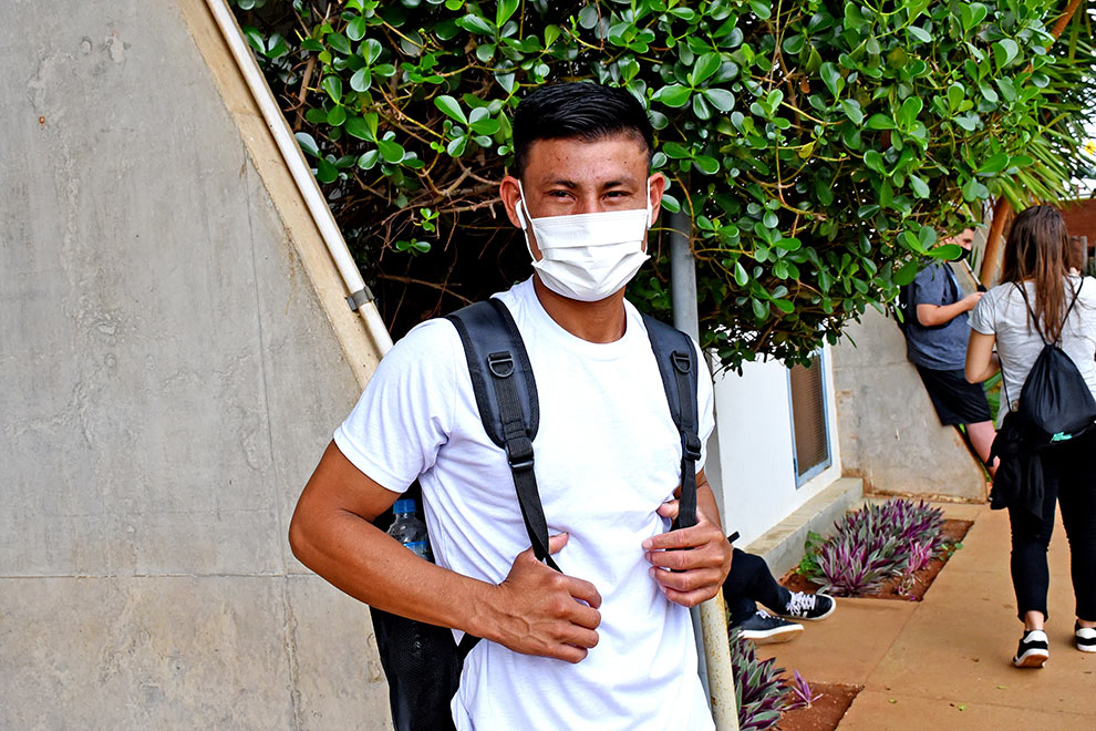 foto mostra estudante. ele veste máscara, camiseta branca e leva mochila nas costas