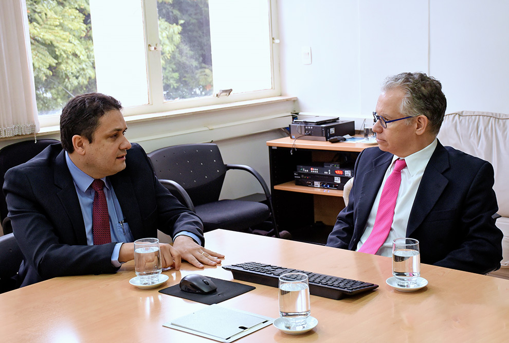 Anderson está sentado em frente ao reitor Marcelo, que escuta com atenção o que o presidente da Capes está falando. Ambos estão vestidos formalmente, com terno e gravada. 