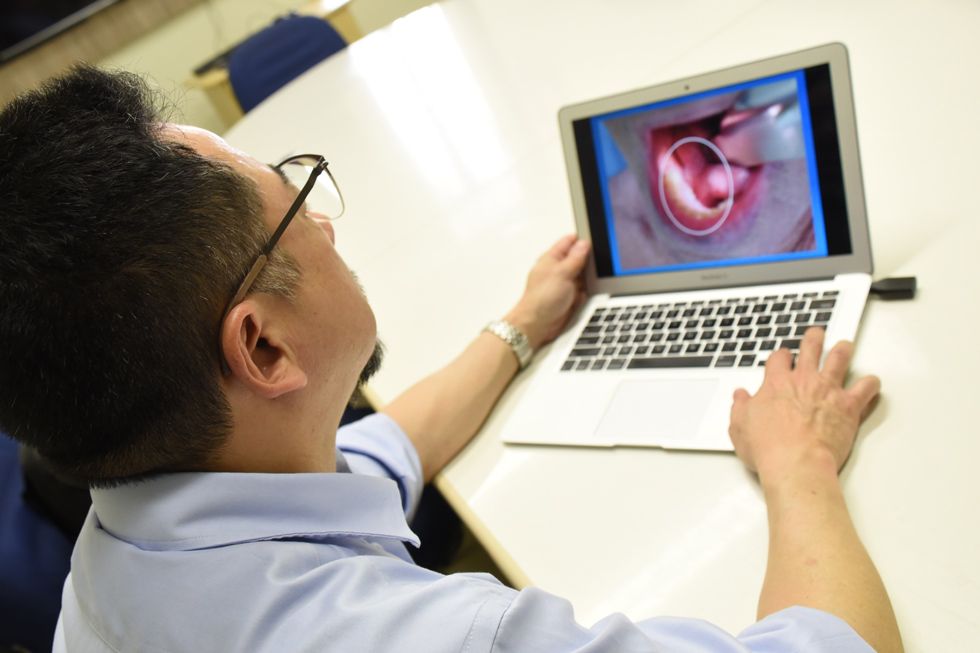 Médico de azul sentado à frente do seu laptop. Ele mostra uma imagem que tem em seu primeiro plano uma boca aberta com um tumor visível