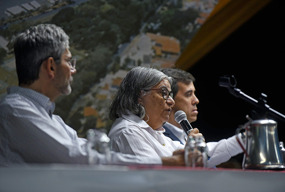 Teresa Atvars com o microfone na mão está sentada entre os outros dois convidados. Apenas seu rosto aparece em destaque.