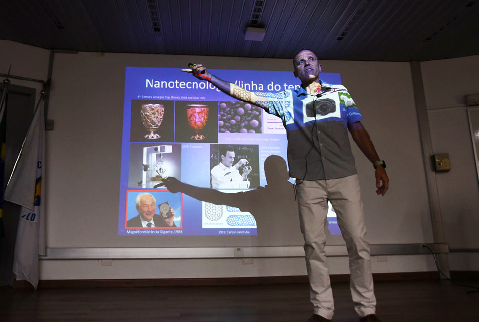 Kleber Pirota é fotografado durante a palestra. Ele está em pé e reflete no corpo uma parte da projeção do telão que está atrás dele. na projeção vemos uma máquina fotográfica na altura do peito do palestrante