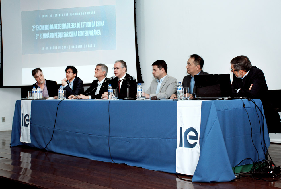 Mesa de abertura do seminário com a presença do reitor. Autoridades estão sentadas e atrás há um telão com o cartaz do seminário