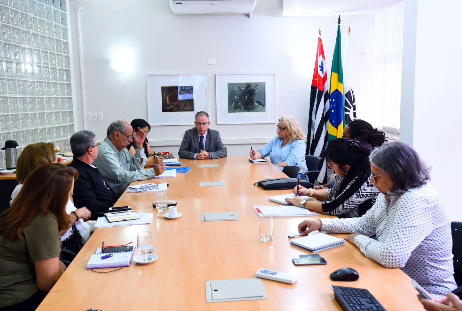 Foto mostra membros da comissão em reunião, sentados à mesa. Na mesa, estão cadernos, blocos, canetas e celulares. Eles olham para o reitor, marcelo knobel, que está na ponta da mesa conduzindo uma apresentação