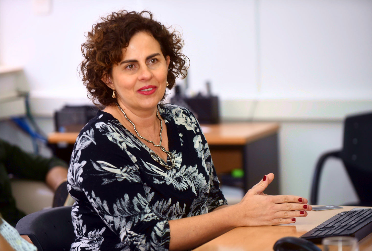 Foto mostra Ana Maria Carneiro enquadrada do colo para cima em uma apresentação. Ela tem cabelos médios, ondulados e veste blusa preta com estampa de folhas brancas