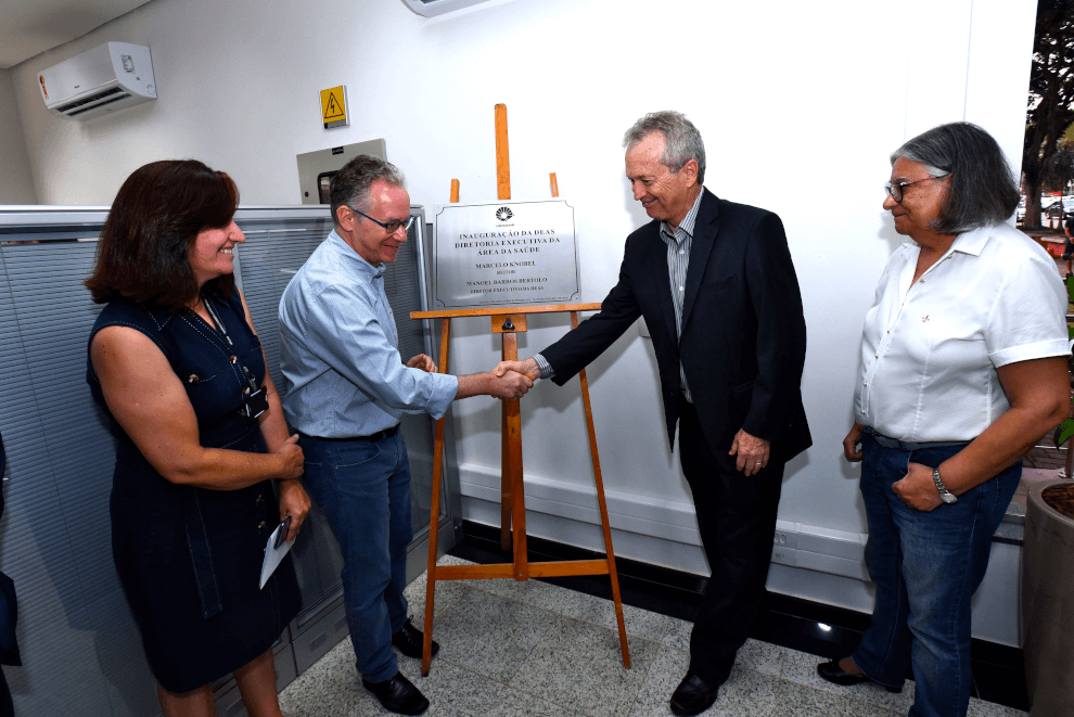 Audiodescrição fotografia colorida mostra o reitor da Unicamp, Marcelo Knobel, a coordenadora geral da Unicamp, teresa Atvars, o diretor executivo da área de saúde, Manoel Bertolo e Sueli Rangel, assessora da diretoria, em frente à placa de inauguração da diretoria executiva da área de saúde. Na foto, o reitor e o diretor executivo estão apertando as mãos. 