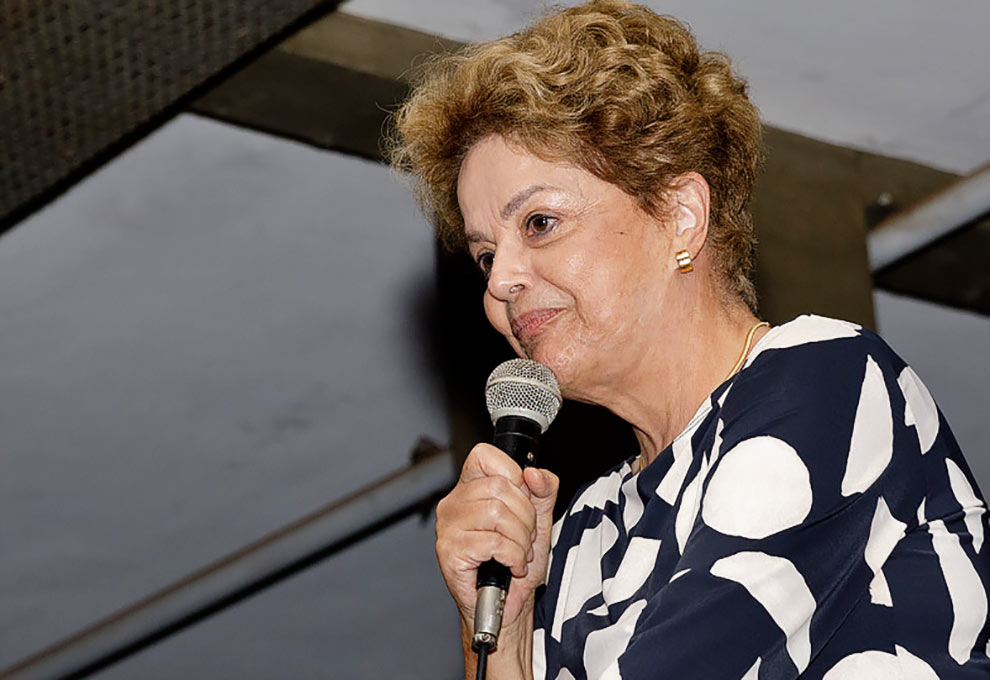 Foto de uma mulher que aparece do peito para cima e de lado. Ela é branca, tem o cabelo castanho e curto. Está usando uma blusa branca e preta e está segurando um microfone.