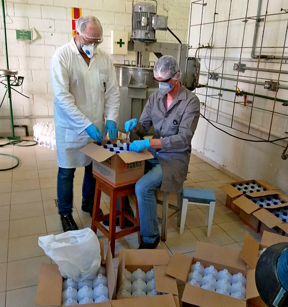 foto mostra dois pesquisadores, vestidos com roupas e máscaras de proteção, mexendo em garrafas plásticas de álcool. atrás está um equipamento de envase do álcool