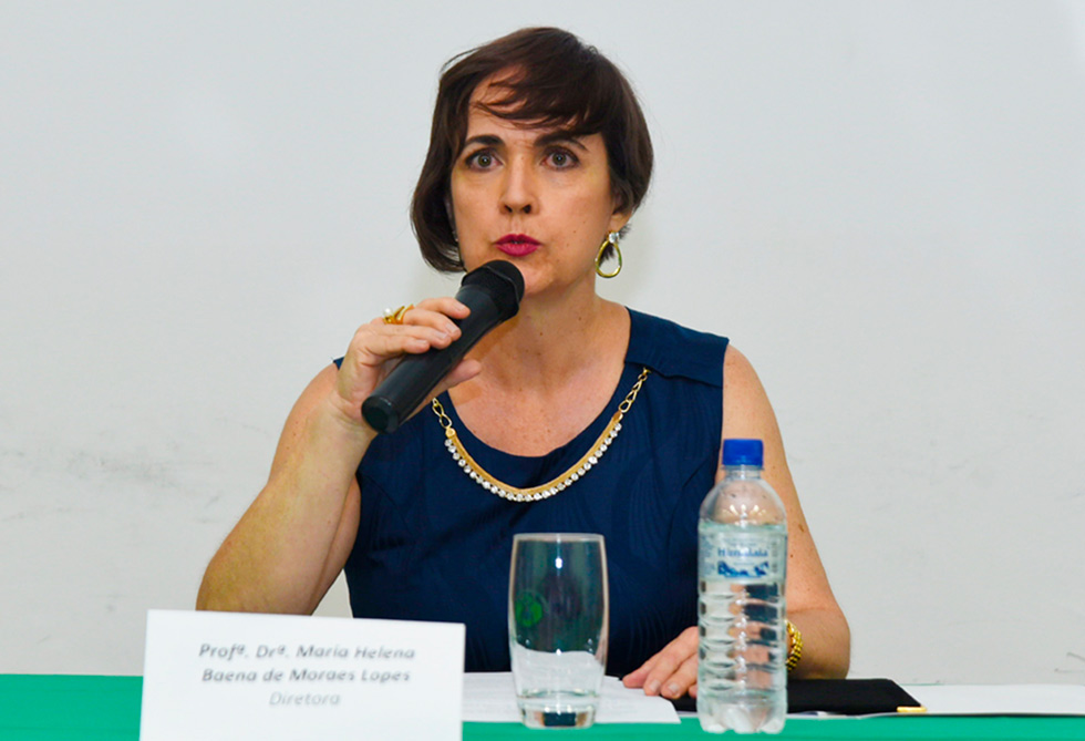 audiodescrição: fotografia colorida da diretora da faculdade de enfermagem da unicamp, maria helena baena. ela está fazendo uma fala, com microfone, e em primeiro plano, na mesa, há uma placa indicando o seu nome