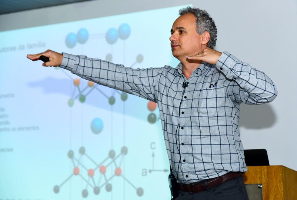 O professor Granado faz um gesto com os braços abertos, o direito mais à frente, para explicar sobre o movimento dos átomos. Atrás dele está um slide de uma apresentação, em tela iluminada, mostrando uma estrutura cristalina