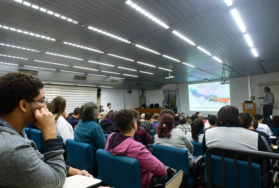 Plateia acompanha a palestra do professor. Imagem mostra as pessoas de costas, sentadas no auditório cheio