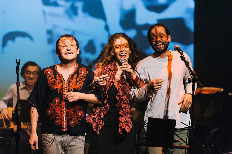 foto mostra, da esquerda para direita, o ator Matheus Nachtergaele, beatriz azevedo e o cantor Moreno Veloso. eles estão cantando no palco com pinturas nos rostos
